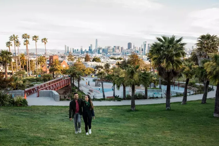 Una coppia cammina verso la telecamera con Dolores Park e lo skyline di 威尼斯人官网平台app dietro di loro.