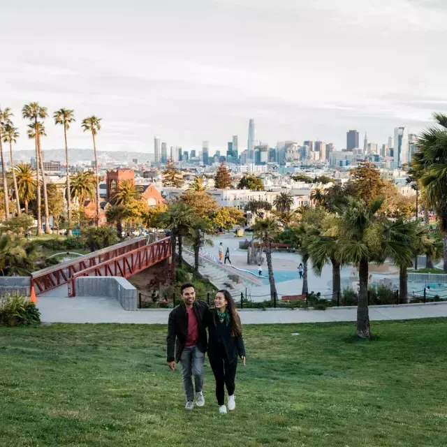 A couple walks toward the camera with Dolores Park and the 威尼斯人官网平台app Skyline behind them.